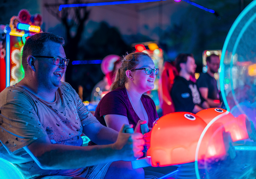 A man and a woman wearing glasses sit side by side on a neon-lit arcade ride, smiling as they grip the handles.