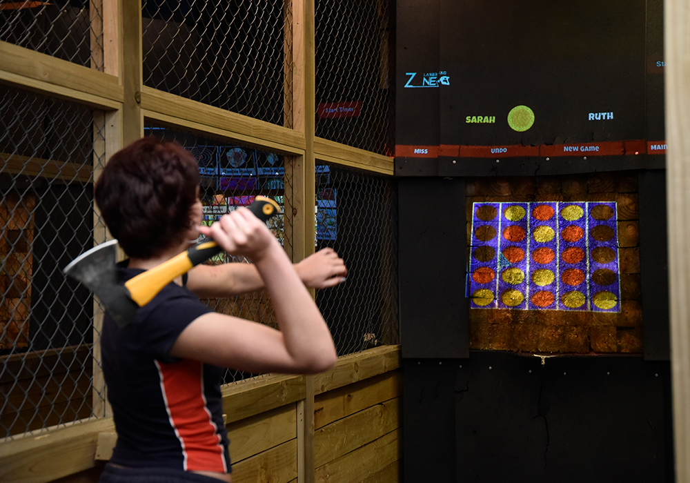 Person in a dark shirt throws an axe at a wooden target in an indoor axe-throwing lane, aiming at a colorful target board.