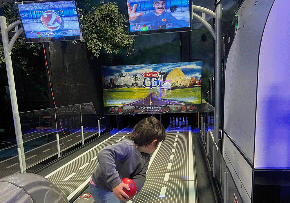 Child in a gray hoodie holding a red bowling ball at an arcade-style bowling lane with screens overhead.