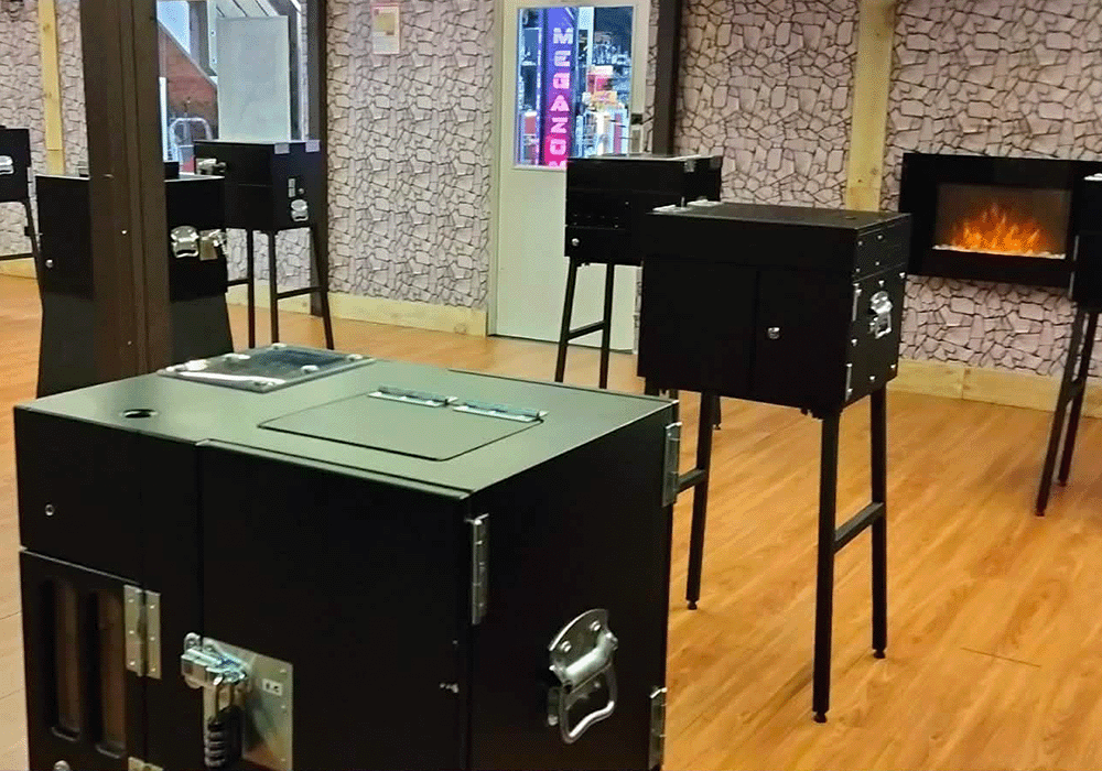 Showroom with multiple black metal stoves on stands arranged around a wooden floor, a fireplace on the wall in the background.