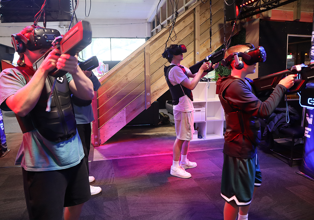 People wearing VR headsets and protective vests, each holding a handheld controller gun in a VR arcade.