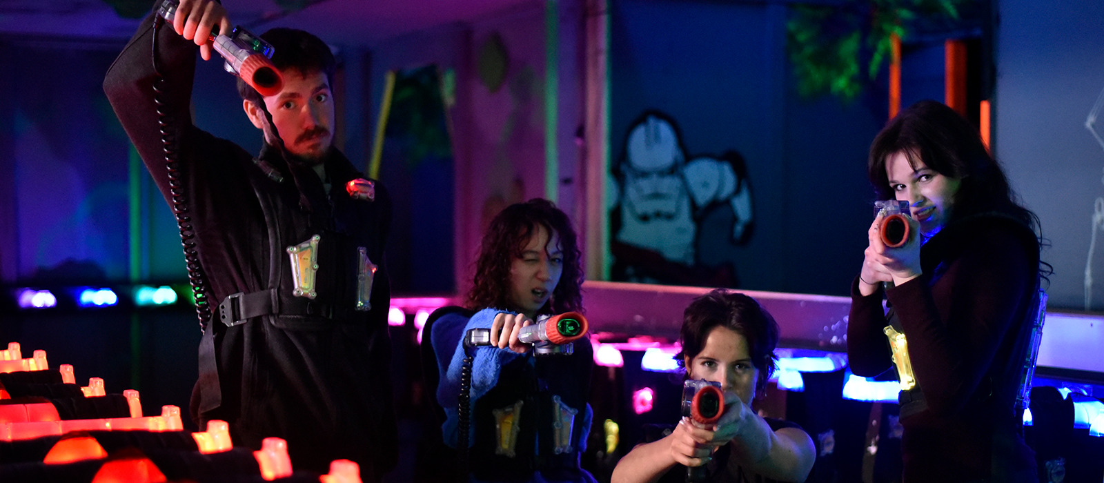 Four players in neon-lit laser tag arena aim laser guns toward the camera.