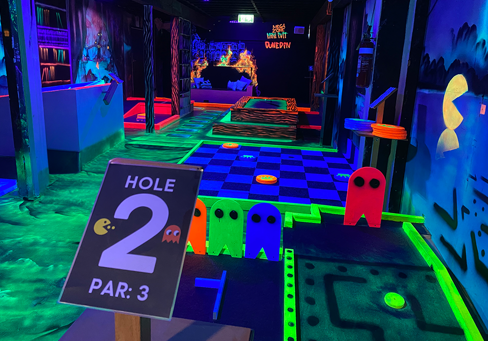 Indoor glow-in-the-dark mini golf with neon Pac-Man ghosts along the edge and a Hole 2, Par 3 sign in foreground