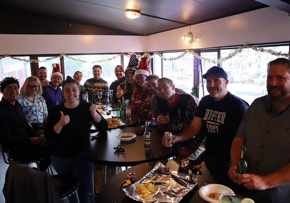 Group of coworkers around a table at a festive holiday party, some wearing Santa hats and smiling at the camera with snacks and drinks on the table.