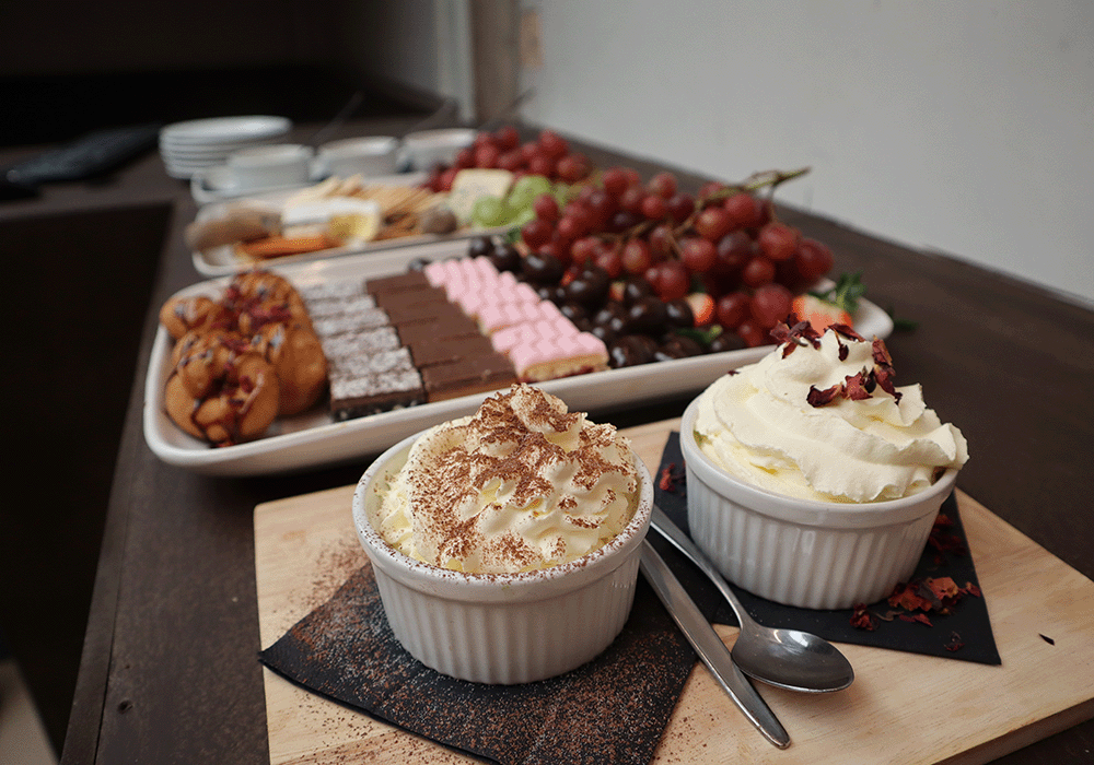 Two whipped-cream desserts in white ramekins on a wooden board, with a large tray of pastries, chocolates, and grapes behind them.