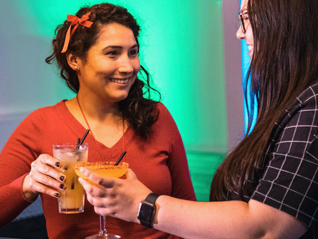 Two women toast with cocktails, smiling at each other in a colorful bar setting.