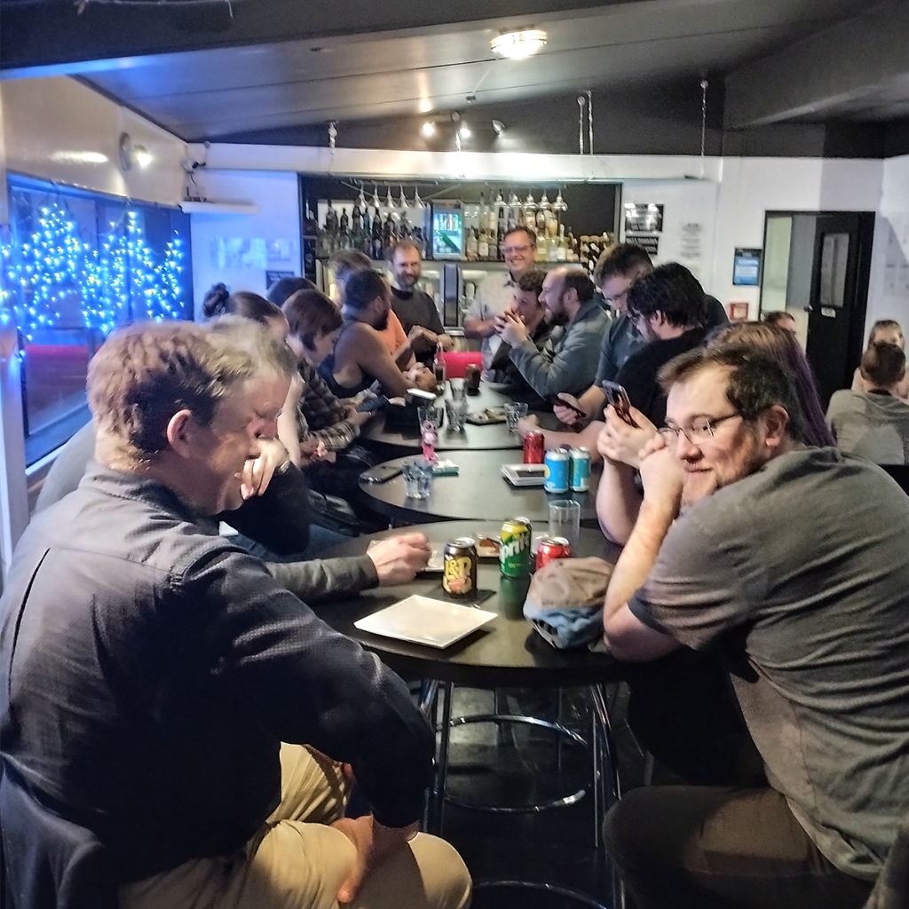 Group of people seated along a long bar table, chatting and drinking in a busy bar setting.