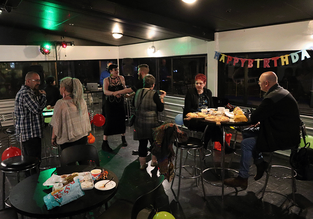 People mingle at a birthday party in a room with a colorful 'Happy Birthday' banner, snacks on tables, and balloons scattered around.