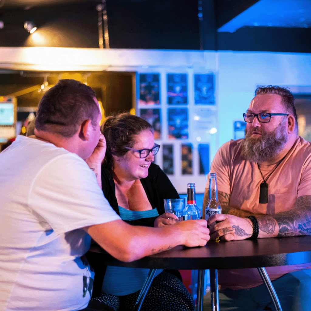 Three adults sit at a bar table, smiling and chatting, with bottles and blue-tinted lighting nearby.